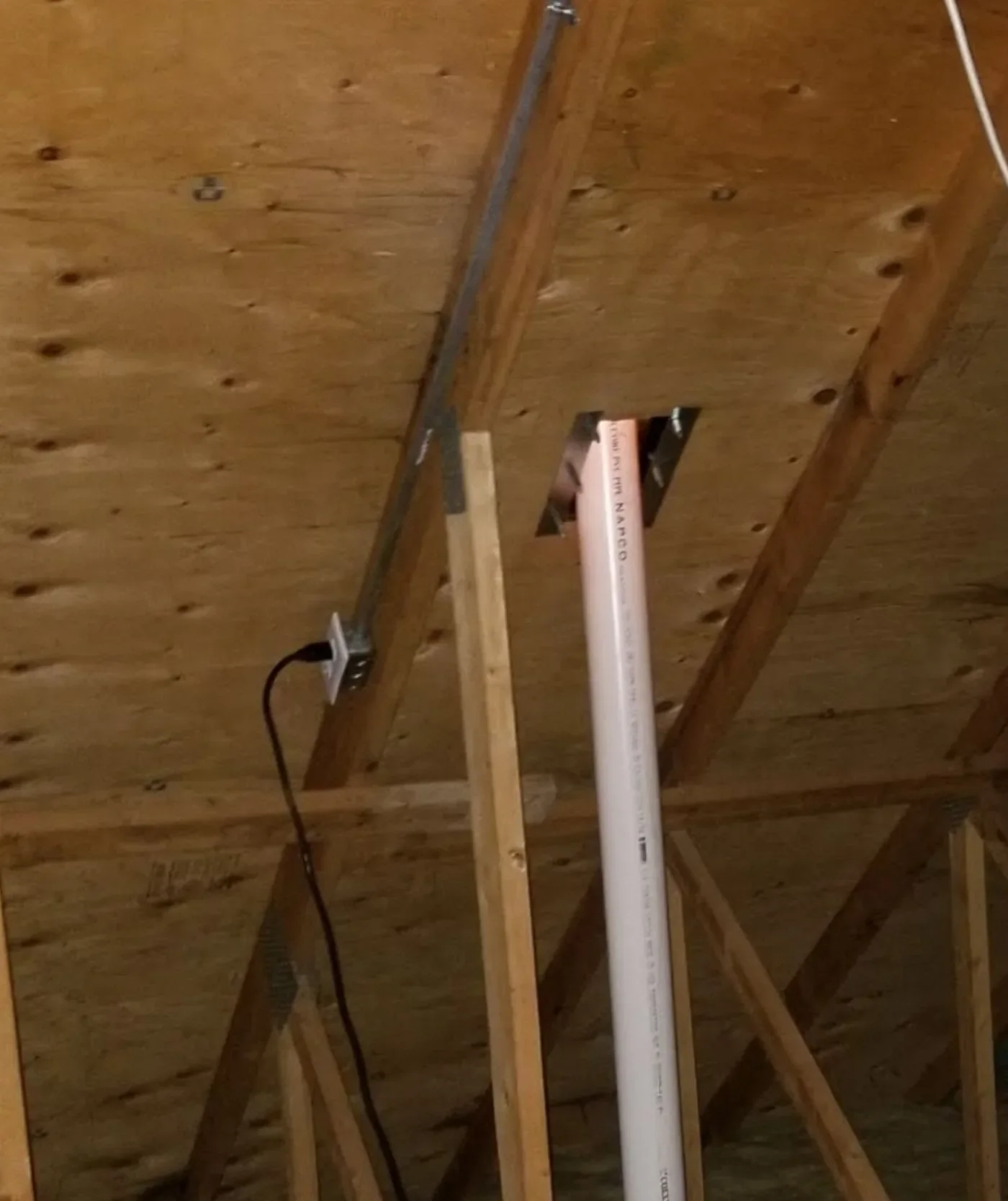 Radon fan unit mounted in an attic above insulation, with PVC pipe exiting through the roof deck.
