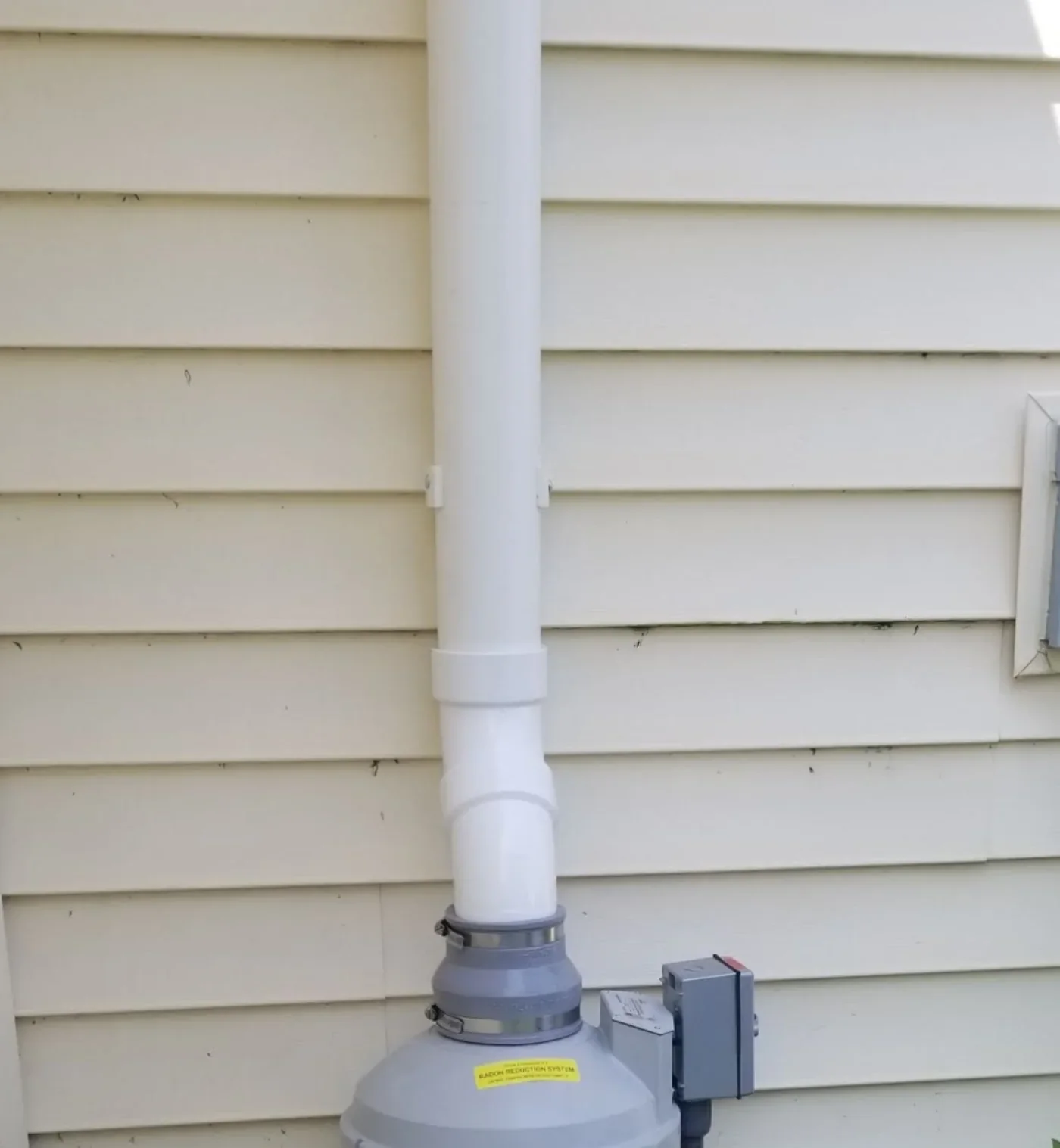 Exterior radon fan unit with a dedicated electrical disconnect box mounted alongside.