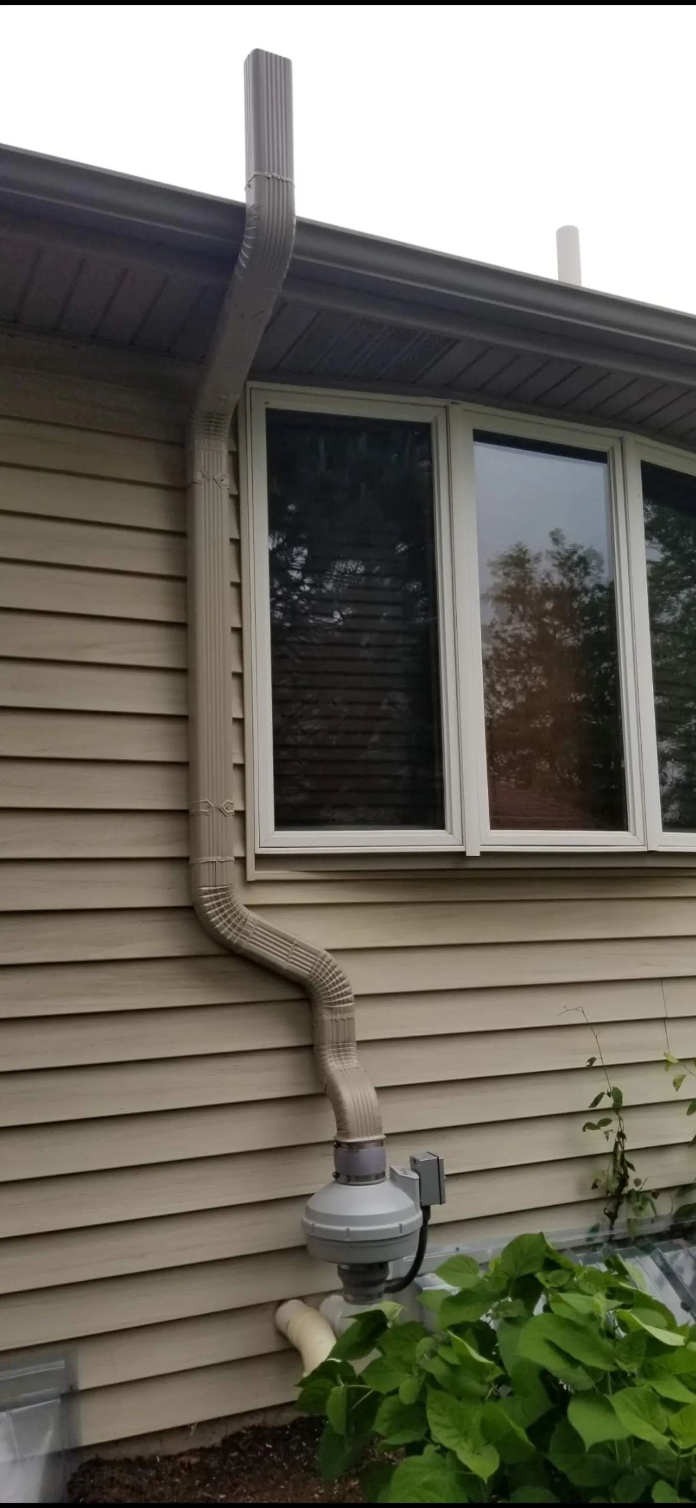 Cream-colored aluminum radon pipe with exterior-mount fan on a tan-sided home.