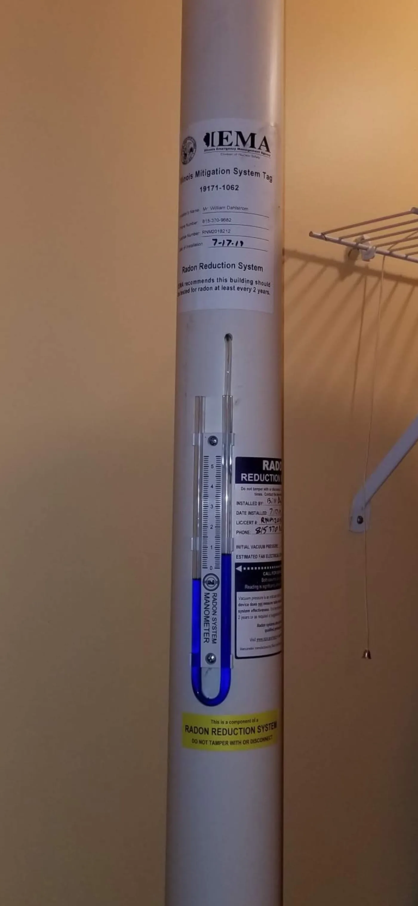 Indoor radon pipe showing a U-tube manometer and the Illinois IEMA Mitigation System Tag.