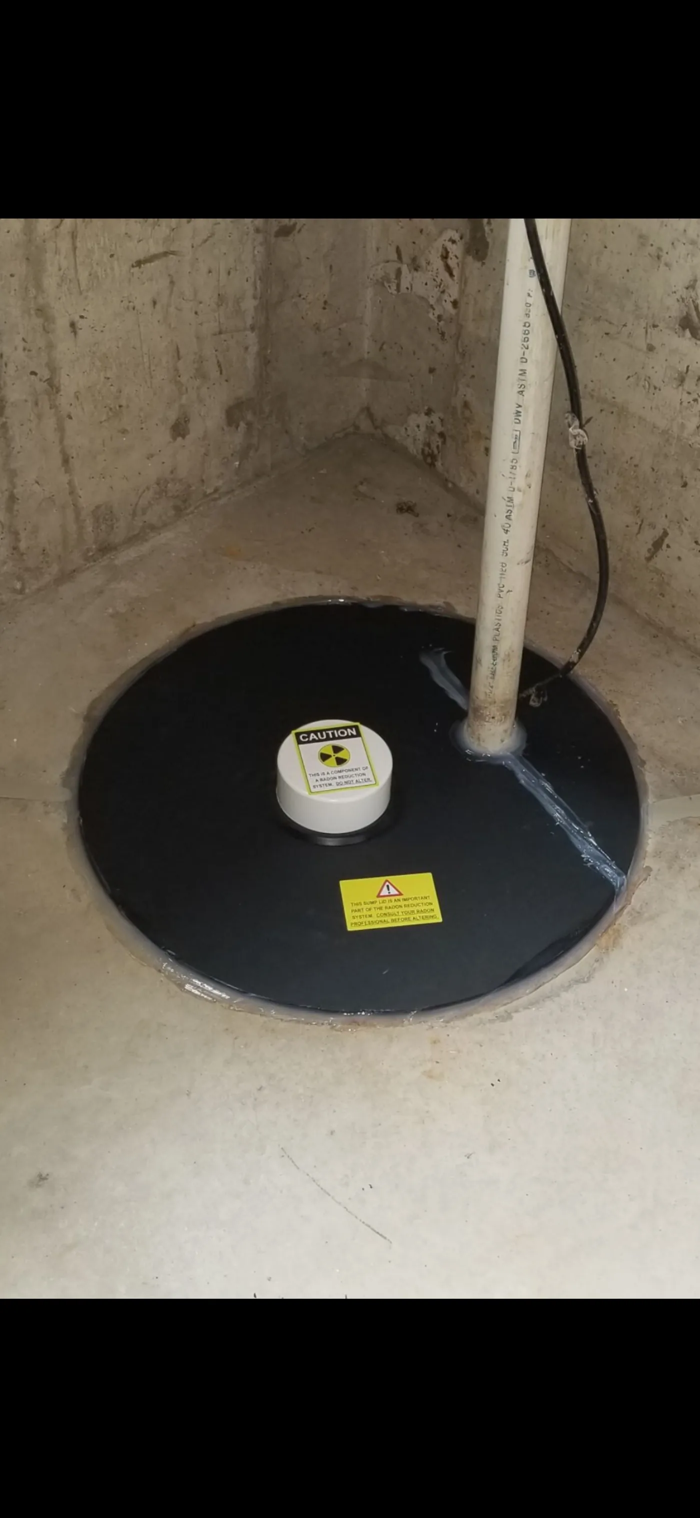 Sealed black sump pit lid with a white radon-system access port and caution labels.